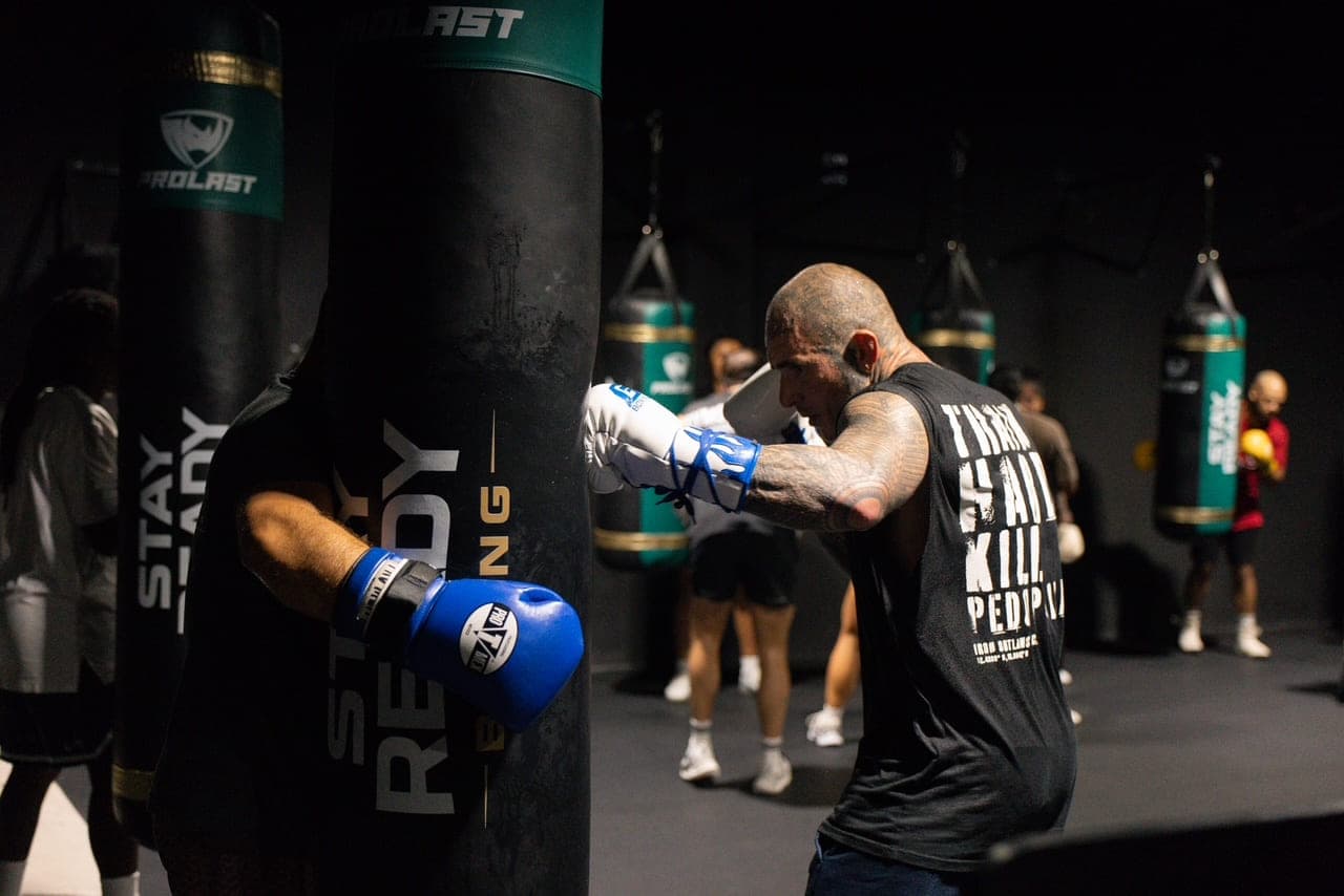 A member in boxing gloves working one of the Stay Ready branded heavy bags, the rest of the bag row receding into the dark behind.
