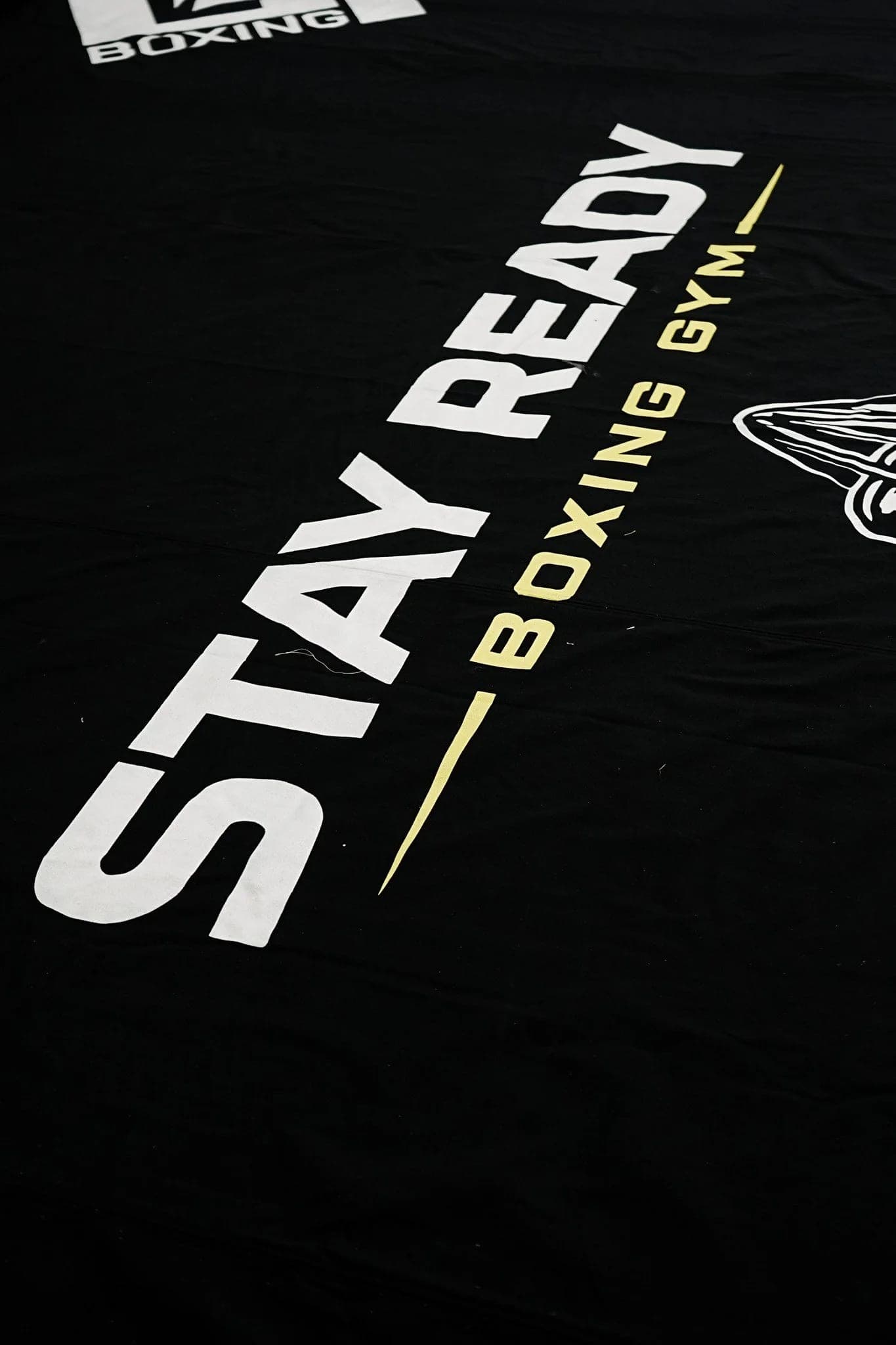 Close detail of the Stay Ready Boxing Gym logo printed on the ring canvas, the fabric creased and worn.