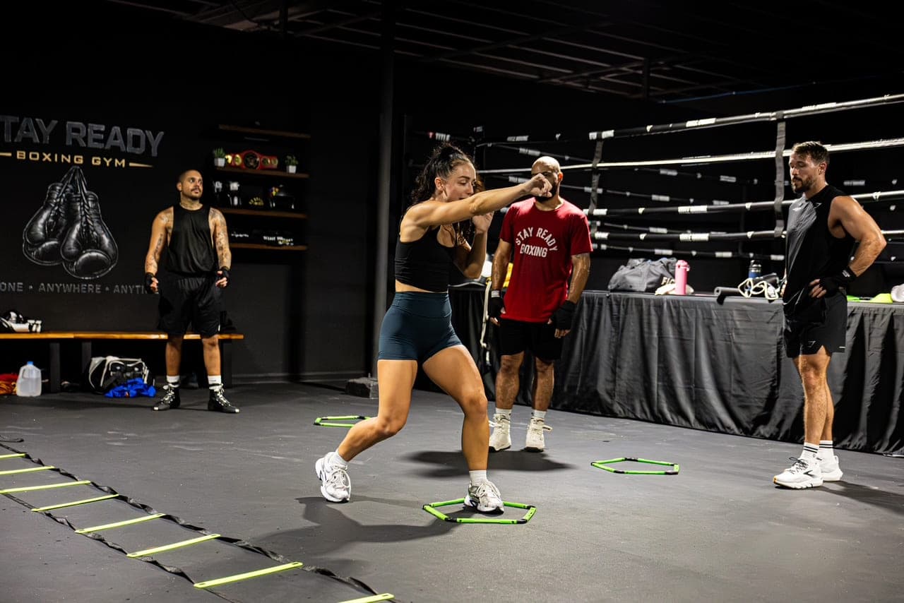 A class in progress across the Stay Ready Boxing Gym floor, one member throwing a punch over agility rings while others wait their turn, the ring at the right and the gym’s glove mural on the wall at the left.