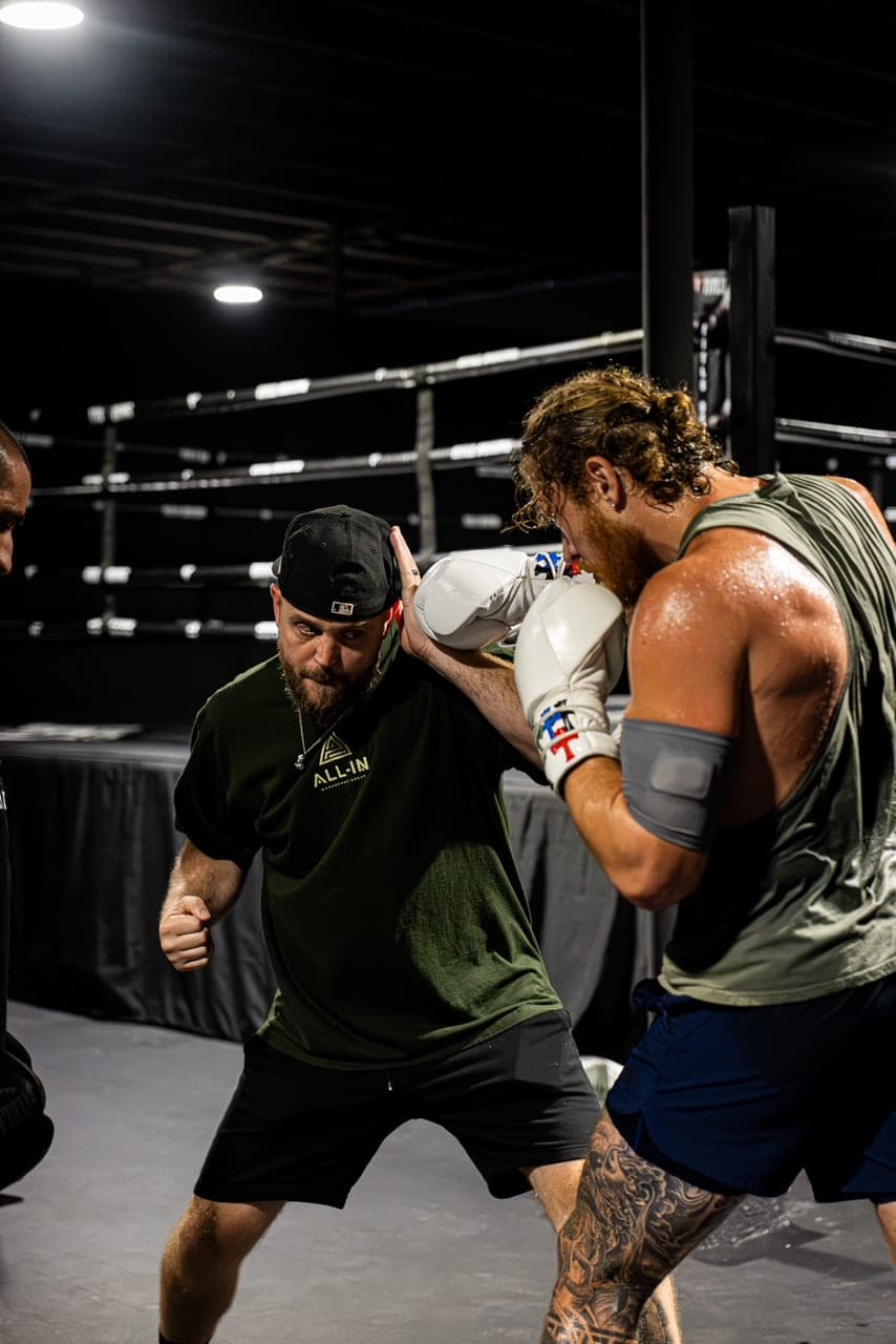 Coach Kevin Gleason working at close range with a boxer in white gloves, the ring behind them.