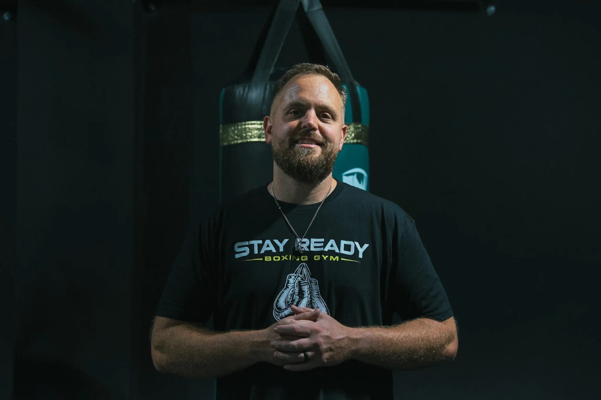 Coach Kevin Gleason in a Stay Ready Boxing Gym shirt, standing in front of a branded heavy bag on the training floor.
