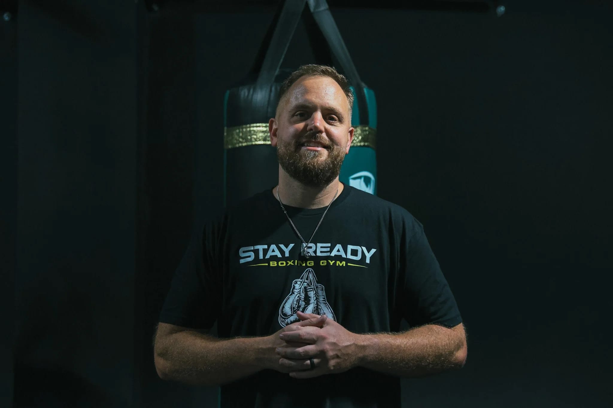 Coach Kevin Gleason in a Stay Ready Boxing Gym shirt, standing in front of a branded heavy bag on the training floor.