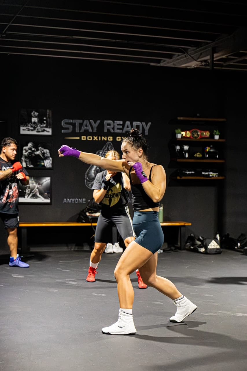 A member in purple hand wraps throwing a straight punch on the Stay Ready Boxing Gym floor, the gym’s name painted on the wall behind and two other people training in the background.