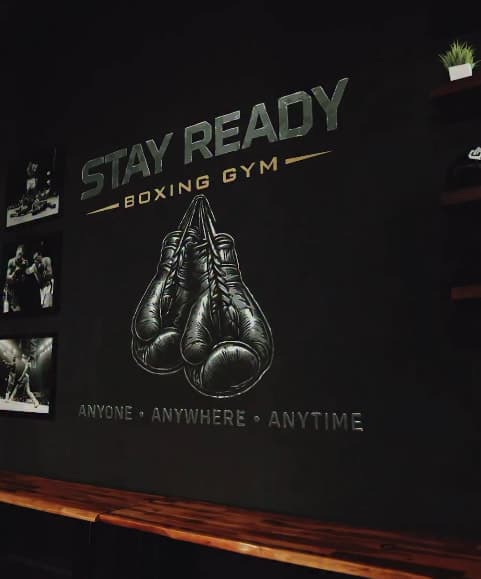 The Stay Ready Boxing Gym mural painted on the training floor wall, the gym’s name and glove mark above the line “Anyone, Anywhere, Anytime”, with framed boxing photographs alongside it.